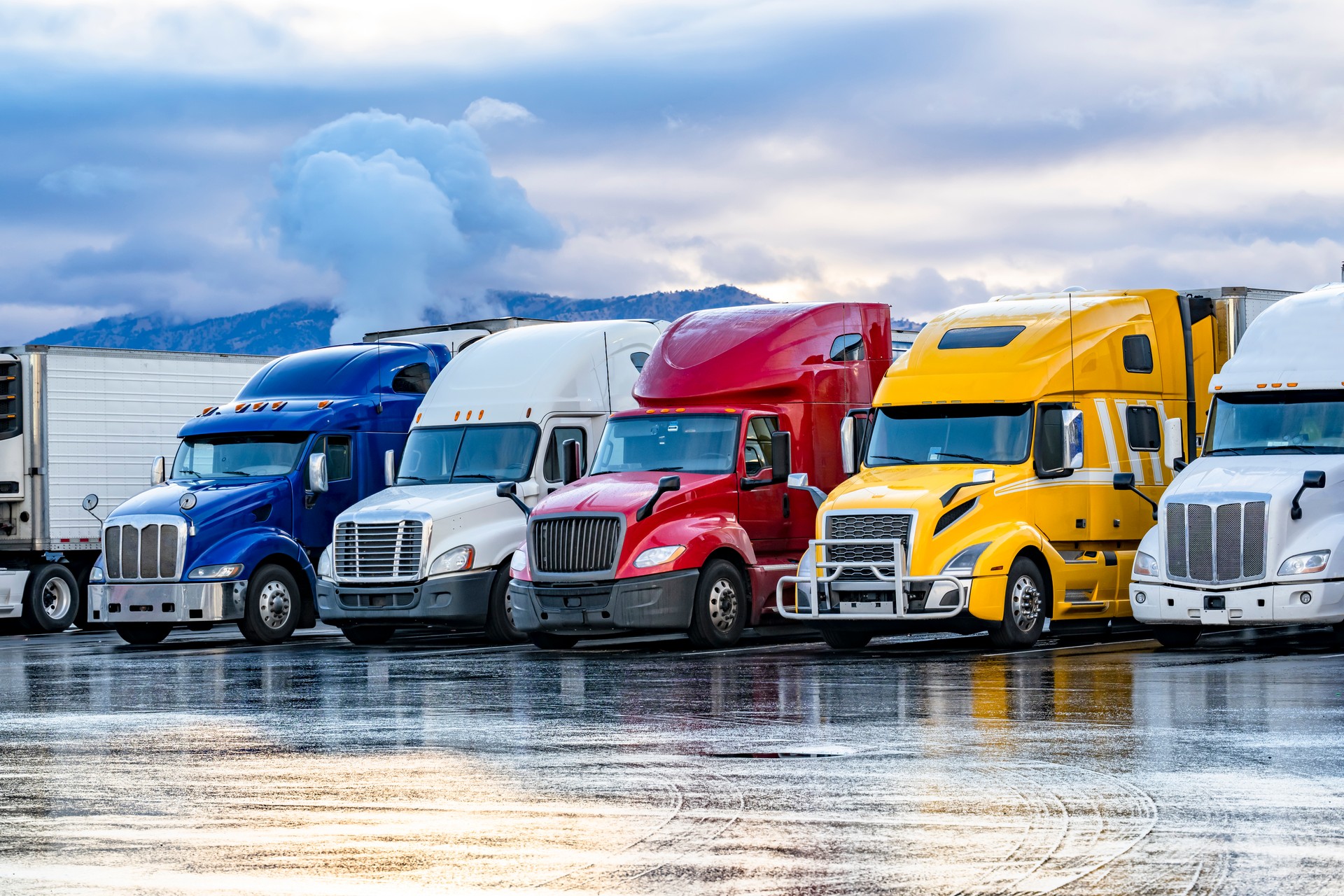 Bright colorful big rigs semi trucks with semi trailers standing in the row on truck stop parking lot at early morning